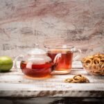 A glass teapot and cup with tea, accompanied by limes and snacks on a rustic wooden table.