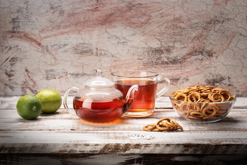 A glass teapot and cup with tea, accompanied by limes and snacks on a rustic wooden table.