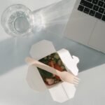 Healthy salad in a takeout box with a glass of water and a laptop on a white surface, top view.